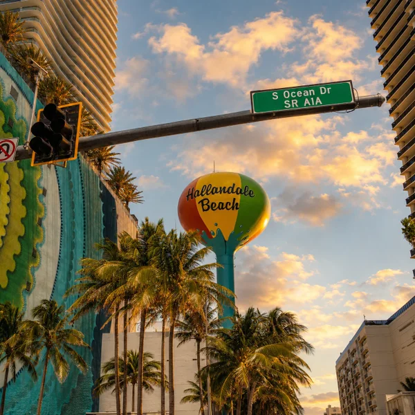 Hallandale Beach, FL sign near Ocean Drive with palm trees, high-rises, and coastal South Florida real estate scenery.