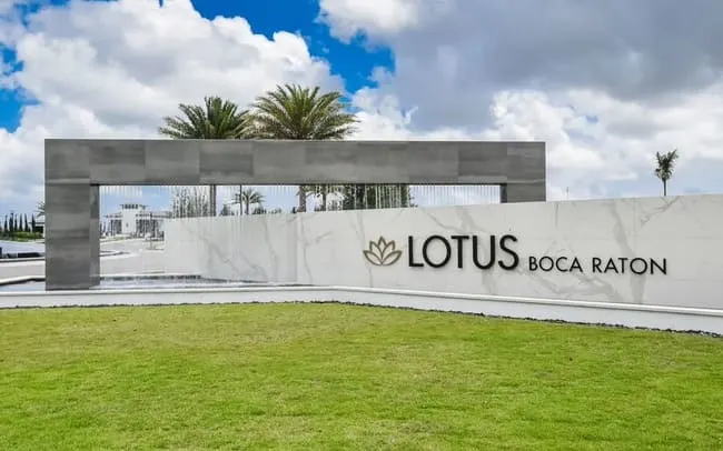 Modern entrance sign at Lotus Boca Raton featuring a marble wall, sleek gray archway, and palm trees under a partly cloudy sky
