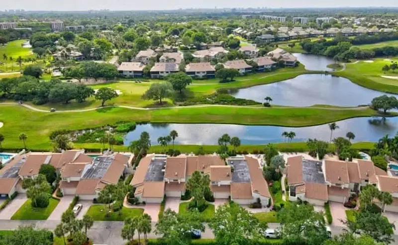 Aerial view of Fairway Oaks Townhomes in Boca West Country Club, Boca Raton, featuring buildings surrounded by greenery with lake and golf view in the background.
