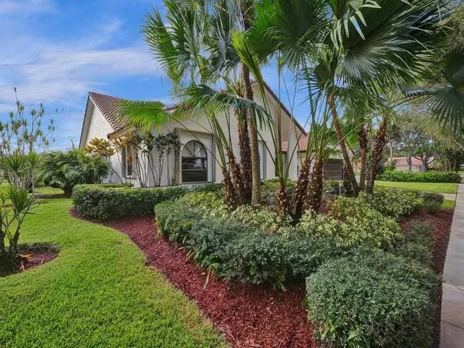 Single-story home with red tile roof, arched window, and landscaped yard featuring palms, hedges, and fresh mulch.