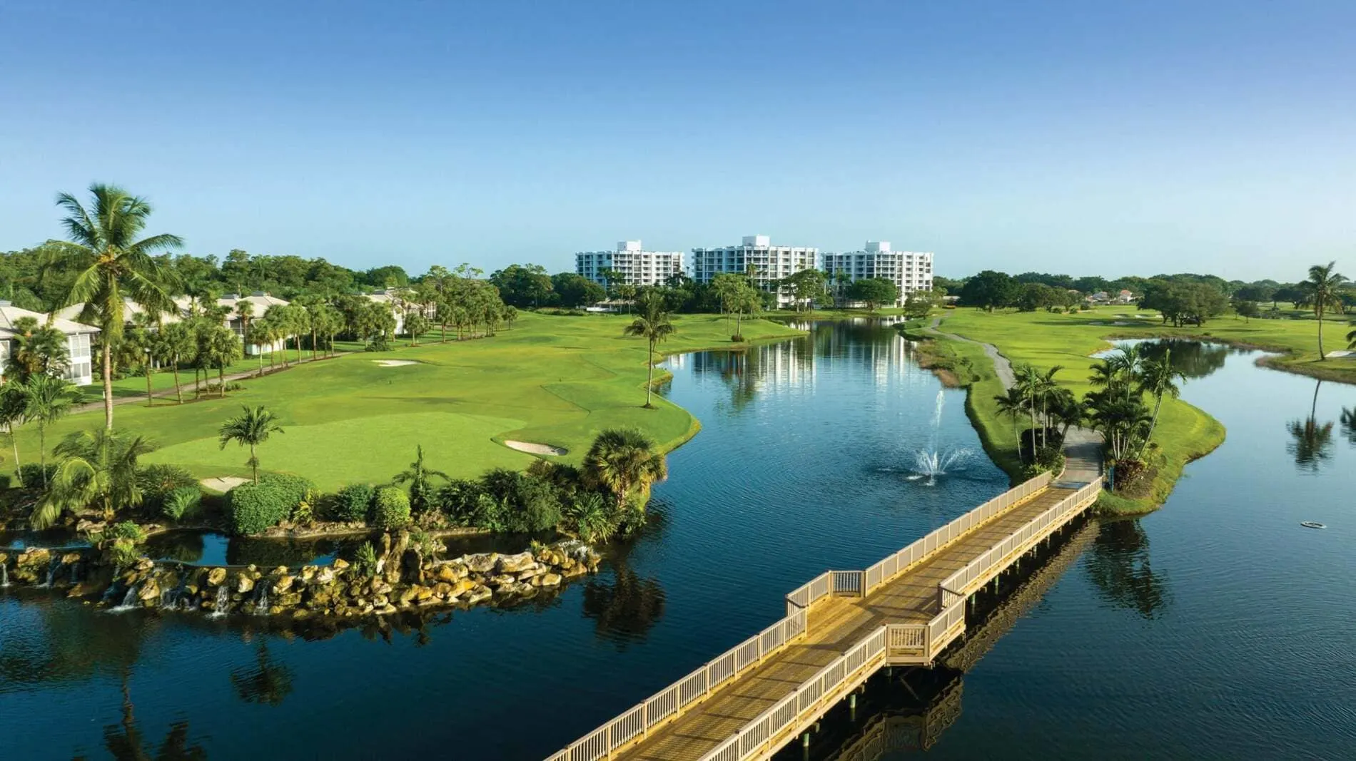 View of Akoya Boca West luxury condos overlooking a lake and golf course with palm trees and walking bridge.
